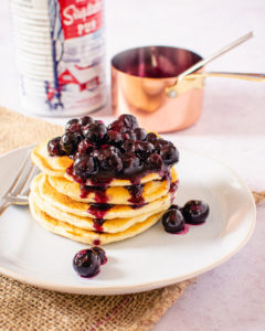 side shot of Blueberry sauce on pancakes staked on a plate with maple syrup